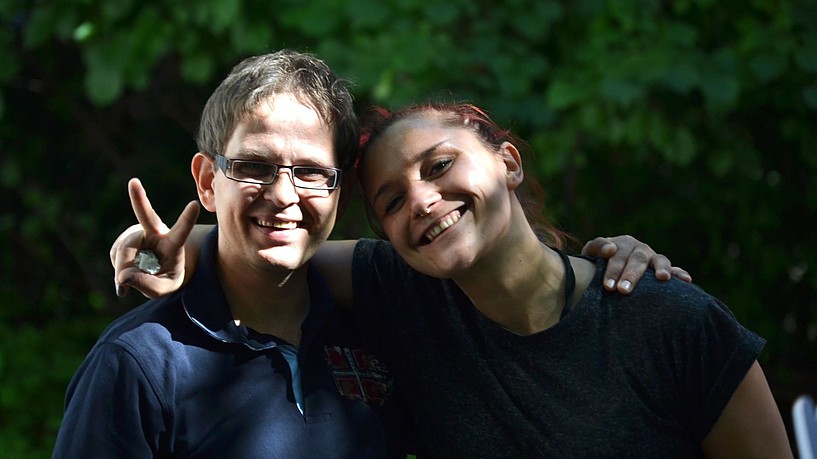 Zwei Personen stehen zusammen und lächeln in die Kamera. Die eine hat den Arm und die andere Person gelegt und macht mit den Fingern ein Peace Zeichen. Im Hintergrund ist ein Teil eines Blätterdaches eines Baumes in tiefem grün zu sehen.